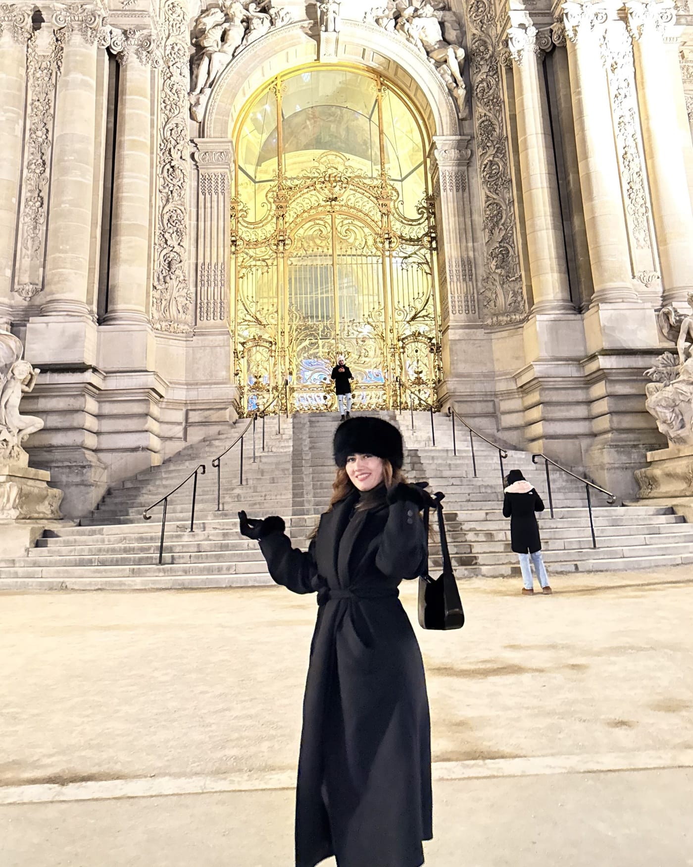 Onesy Carranza con abrigo largo negro y gorro de pelo frente al Petit Palais de París iluminado de noche