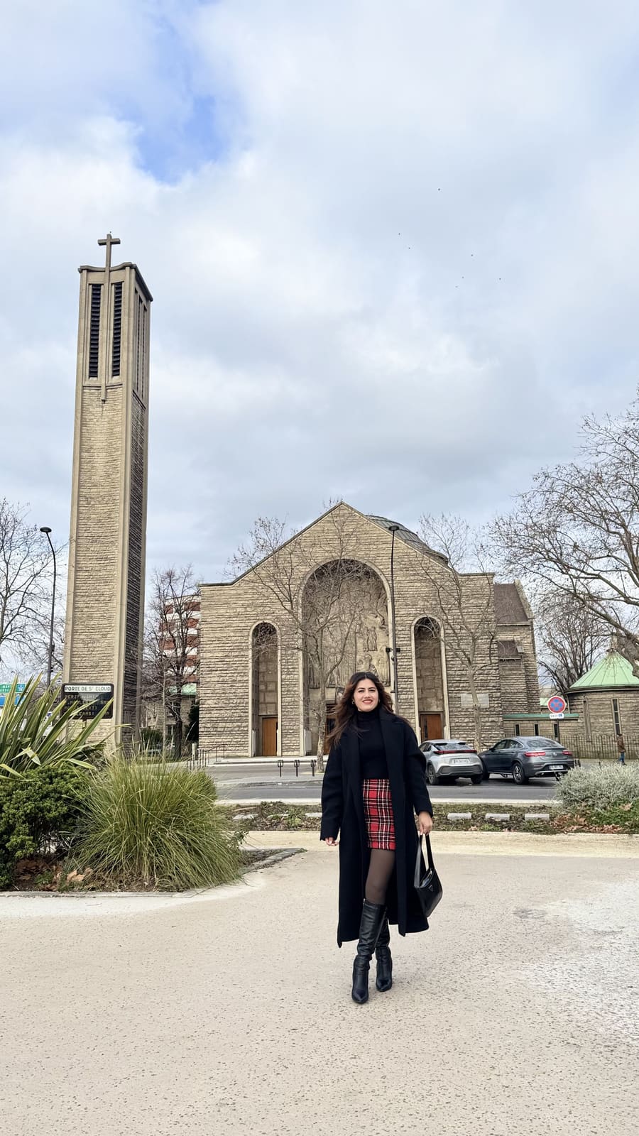 Onesy Carranza con abrigo largo negro, falda de cuadros y botas altas frente a una iglesia de piedra